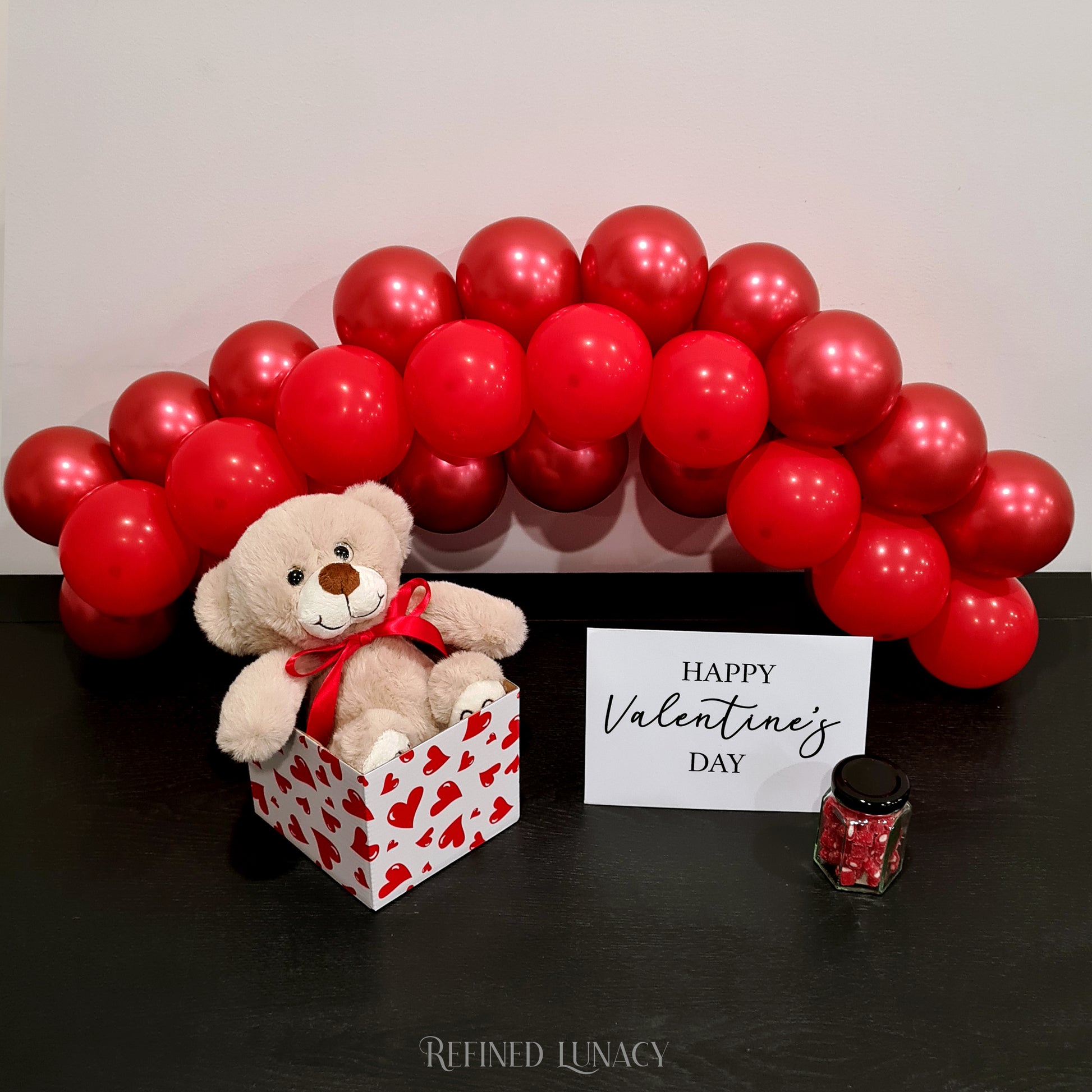 Red balloon garland with plush teddy bear in heart box with Happy Valentine's Day card and heart rock candy