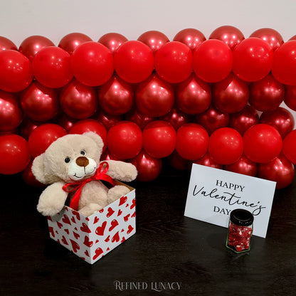 Red balloon garland with plush teddy bear in heart box with Happy Valentine's Day card and heart rock candy