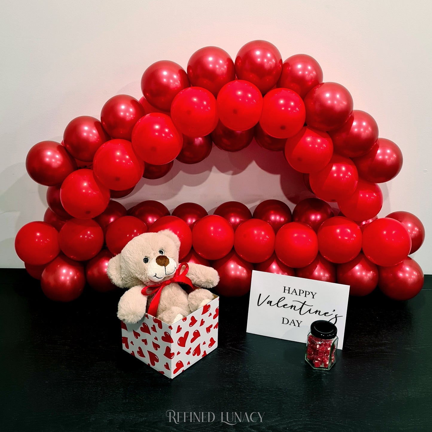 Red balloon garland with plush teddy bear in heart box with Happy Valentine's Day card and heart rock candy